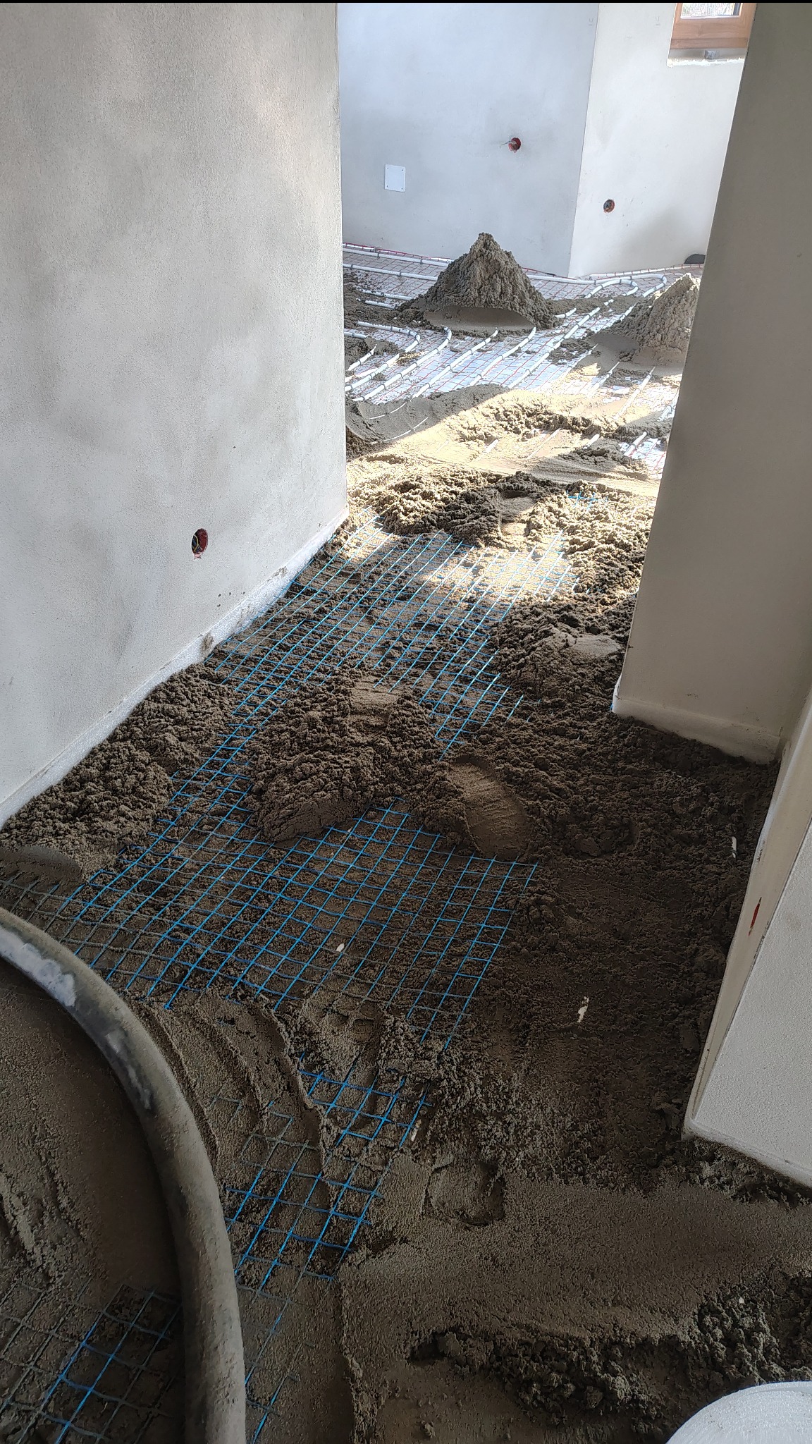 Wnętrze w trakcie prac: na podłodze siatka zbrojeniowa, częściowo pokryta mokrą wylewką. W tle widoczne kopce materiału i rury ogrzewania podłogowego.