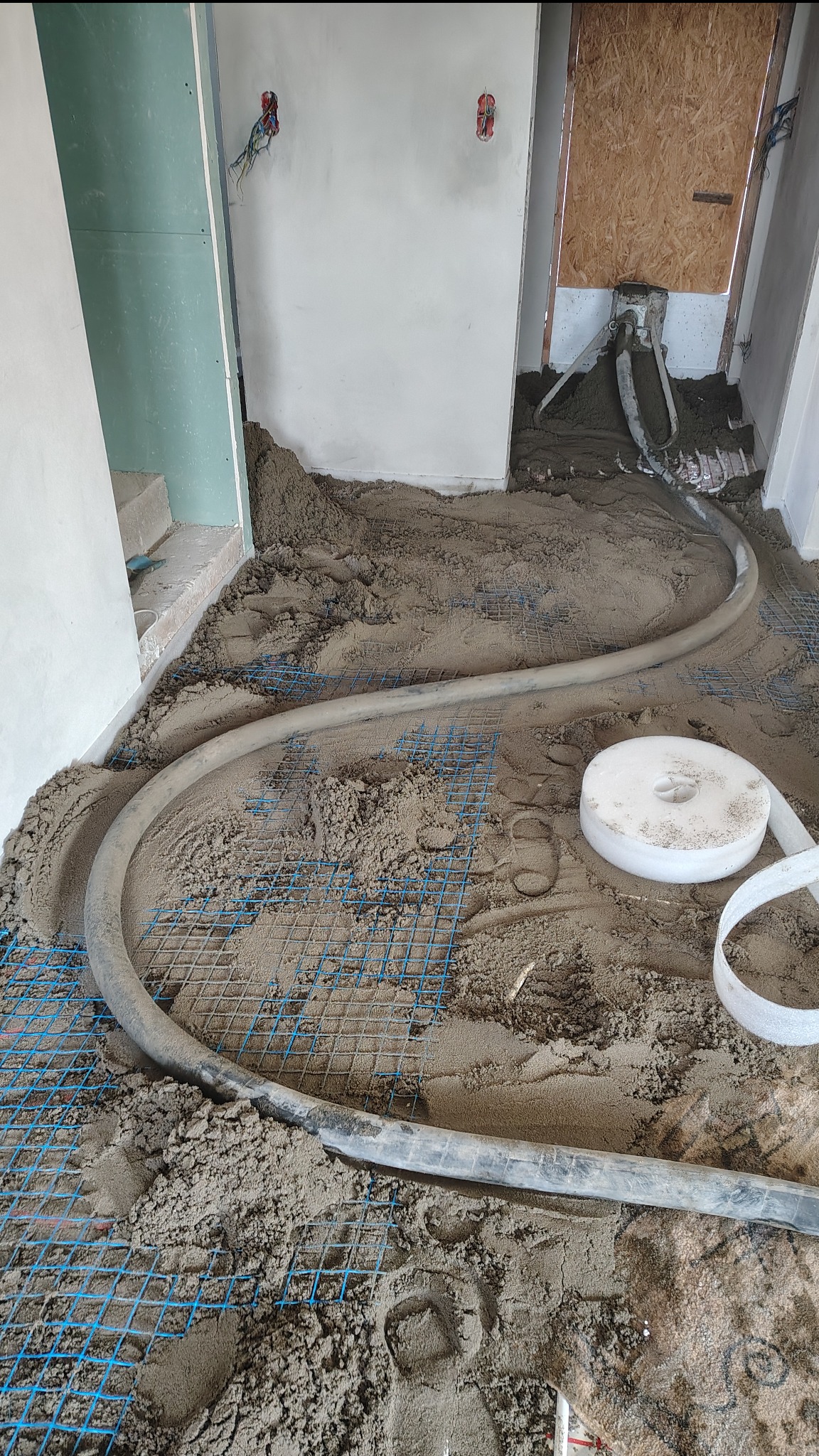 Wylewka jastrychowa w trakcie realizacji: szara masa na siatce z rurą doprowadzającą, widoczne ślady stóp. Surowe ściany i instalacje elektryczne w tle.