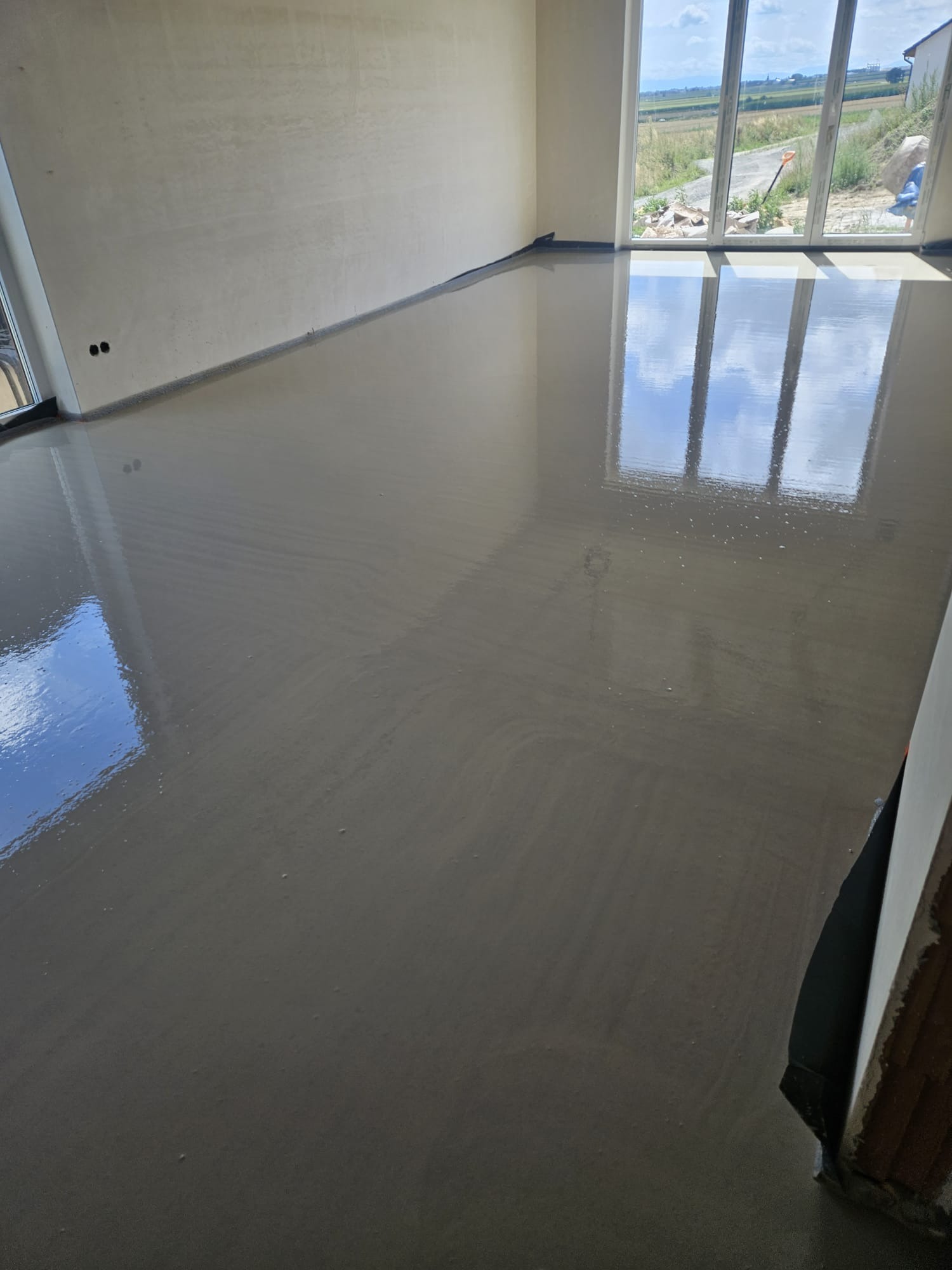 Świeżo wylana, gładka posadzka w jasnym pomieszczeniu z dużymi oknami, odbijającymi niebo. Widoczne ślady po rozprowadzaniu masy, podkreślające ręczne wykonanie.