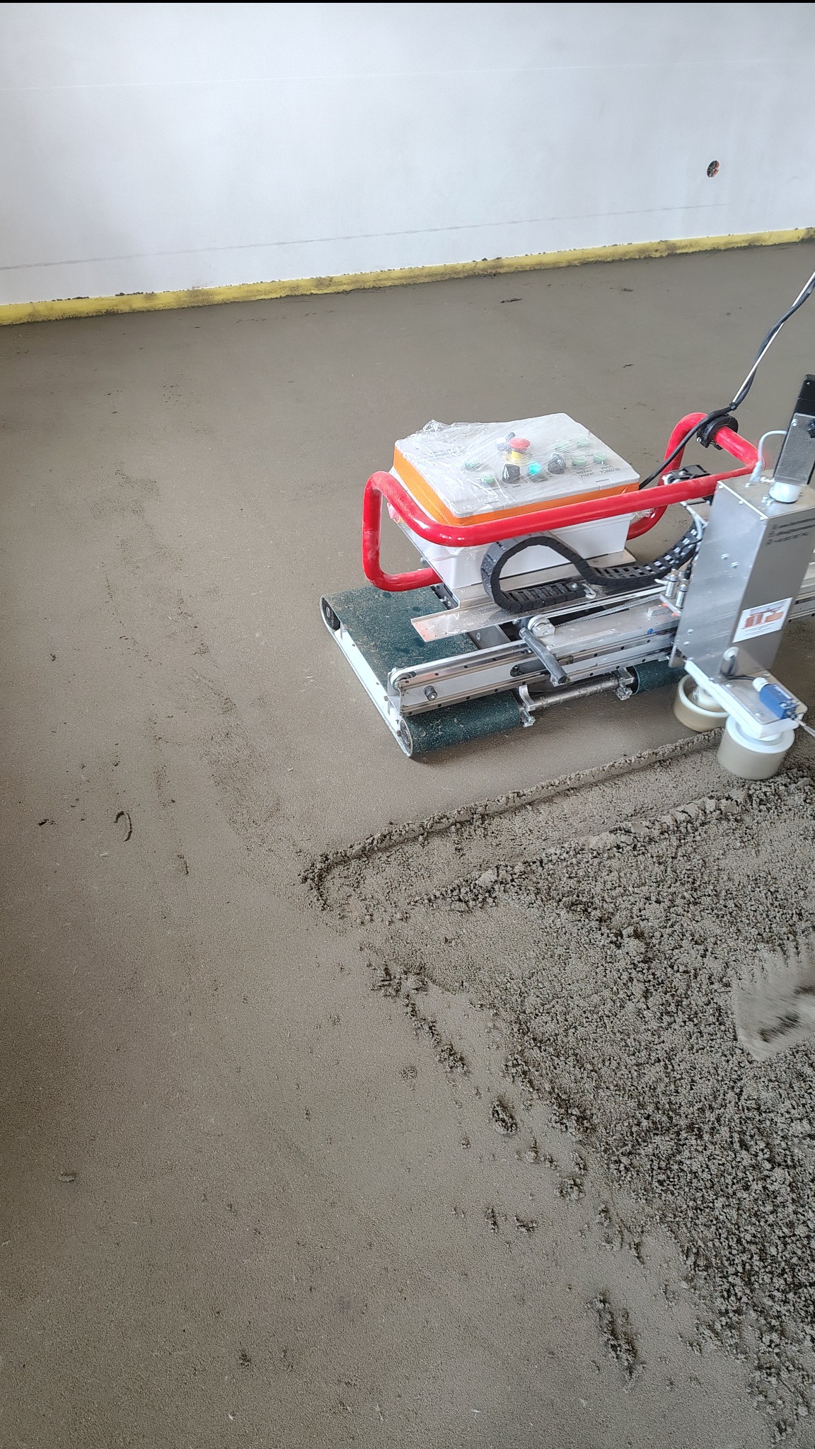 Praca maszyny do wylewania posadzki w Lipiu. Widoczne ślady po pracy urządzenia na świeżej wylewce. Żółta taśma przy ścianie.