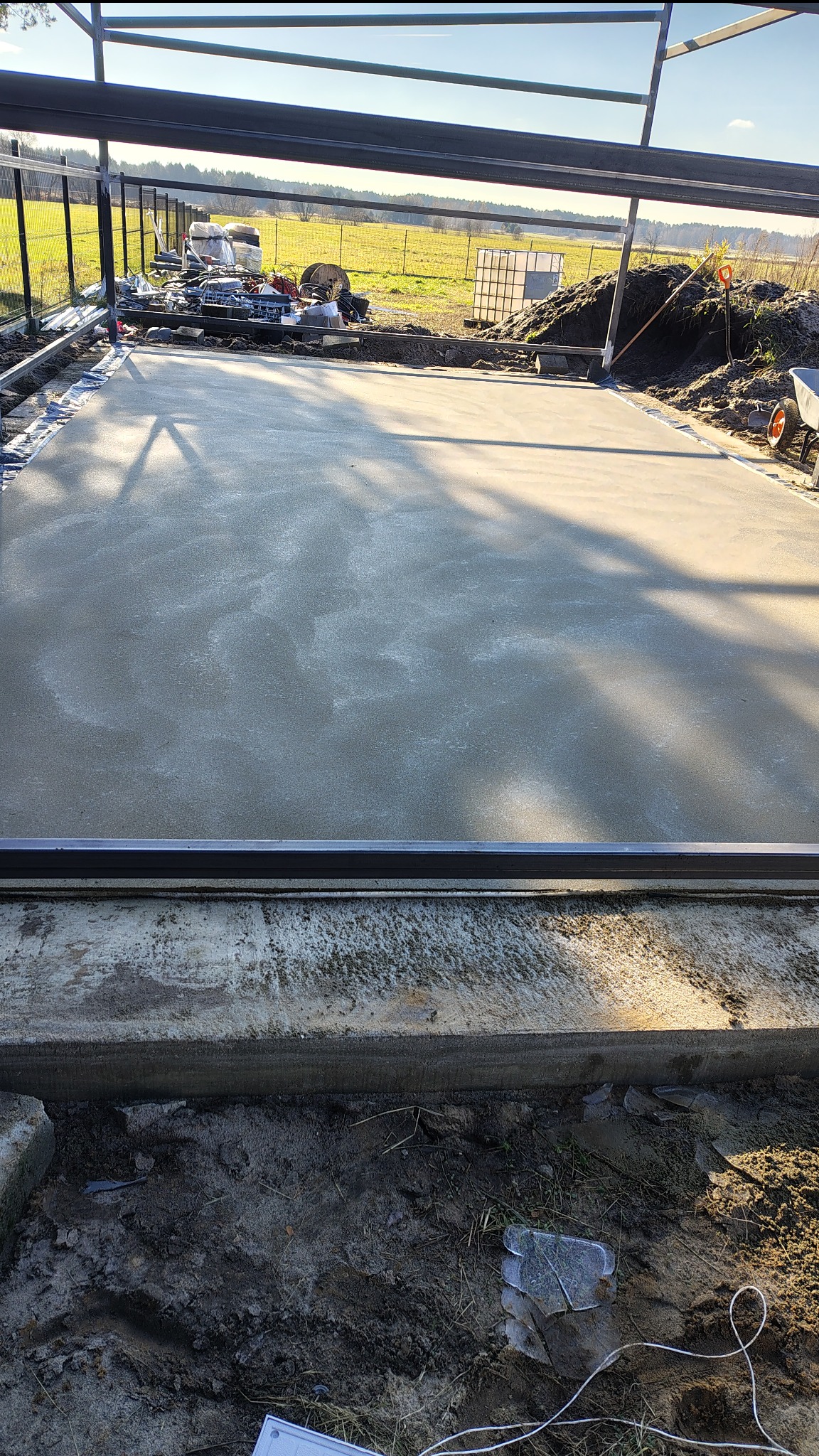 Świeżo wylana, gładka posadzka na zewnątrz budynku w trakcie budowy, z widocznym zbrojeniem i elementami konstrukcyjnymi. W tle krajobraz polny.