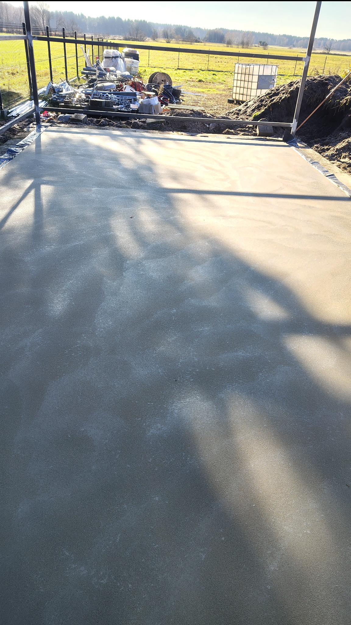 Świeżo wylana, gładka posadzka na zewnątrz, w tle widoczne ogrodzenie i teren zielony. Na posadzce widoczne cienie drzew i odbicia światła słonecznego.