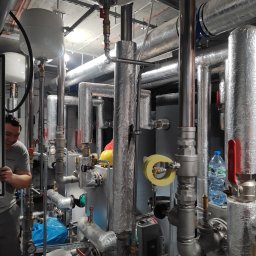 Volodymyr Horokhovets HorTech Energy - Złożona instalacja grzewcza z rurami w izolacji, zbiornikami wyrównawczymi i zaworami. Widoczny fragment osoby. Elementy instalacji podwieszone pod sufitem.