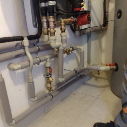 Volodymyr Horokhovets HorTech Energy - Złożona instalacja grzewcza z rurami w izolacji, zaworami i pompą obiegową, zamontowana przy ścianie i zbiorniku. Widoczny fragment osoby w dolnym rogu.