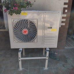 Volodymyr Horokhovets HorTech Energy - Pompa ciepła powietrze-woda na zewnątrz budynku, zamontowana na metalowym stelażu, z wiszącą doniczką z kwiatami nad nią. Widoczne płytki na podłożu.