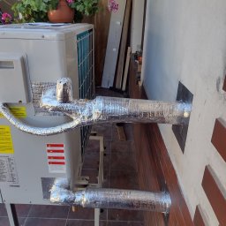 Volodymyr Horokhovets HorTech Energy - Zewnętrzna jednostka klimatyzacji z izolowanymi rurami, przymocowanymi do ściany budynku. Na górze jednostki doniczka z kwitnącymi kwiatami.