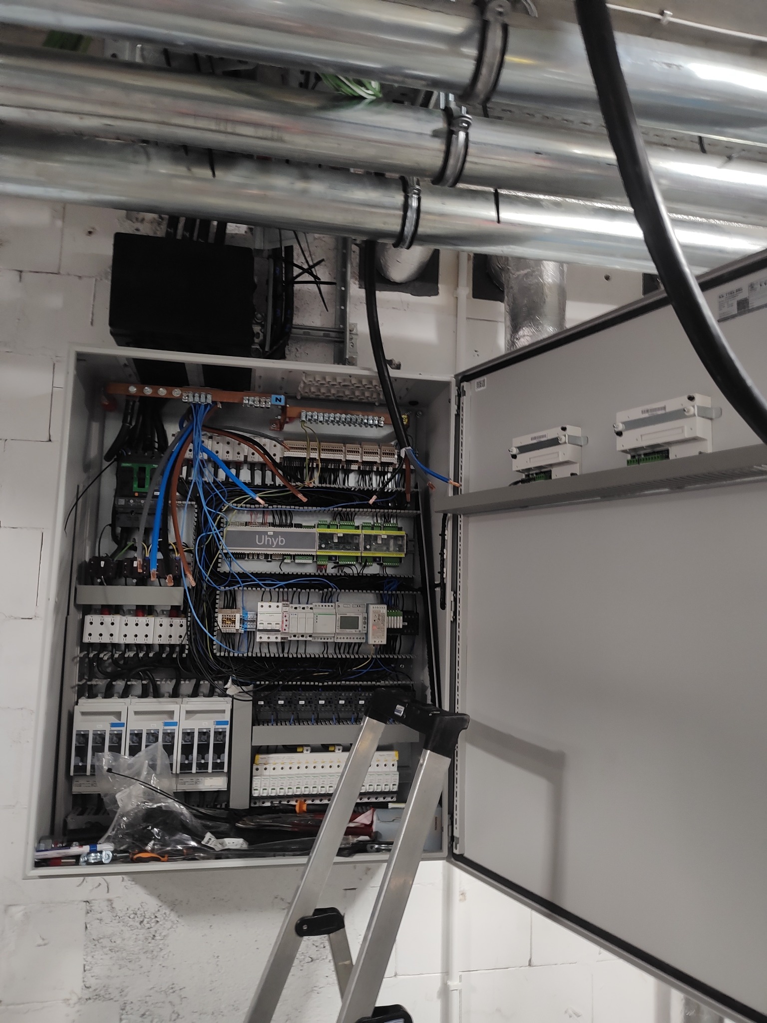 Otwarta skrzynka elektryczna z widocznymi przewodami, bezpiecznikami i elementami sterującymi. Nad skrzynką rury instalacyjne. U dołu drabina.