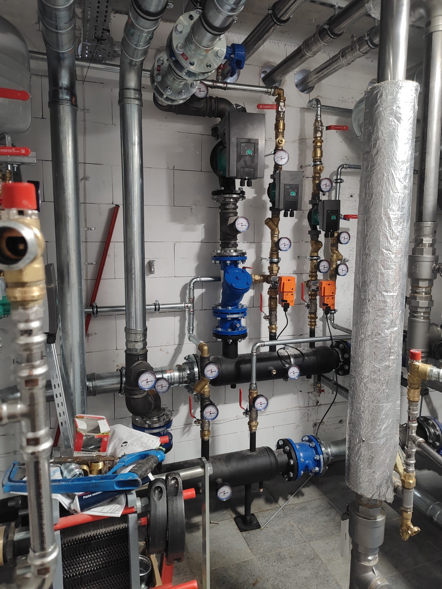 Złożona instalacja grzewcza z rurami, zaworami i manometrami na tle białych płytek. Widoczne elementy izolacji termicznej i narzędzia hydrauliczne.