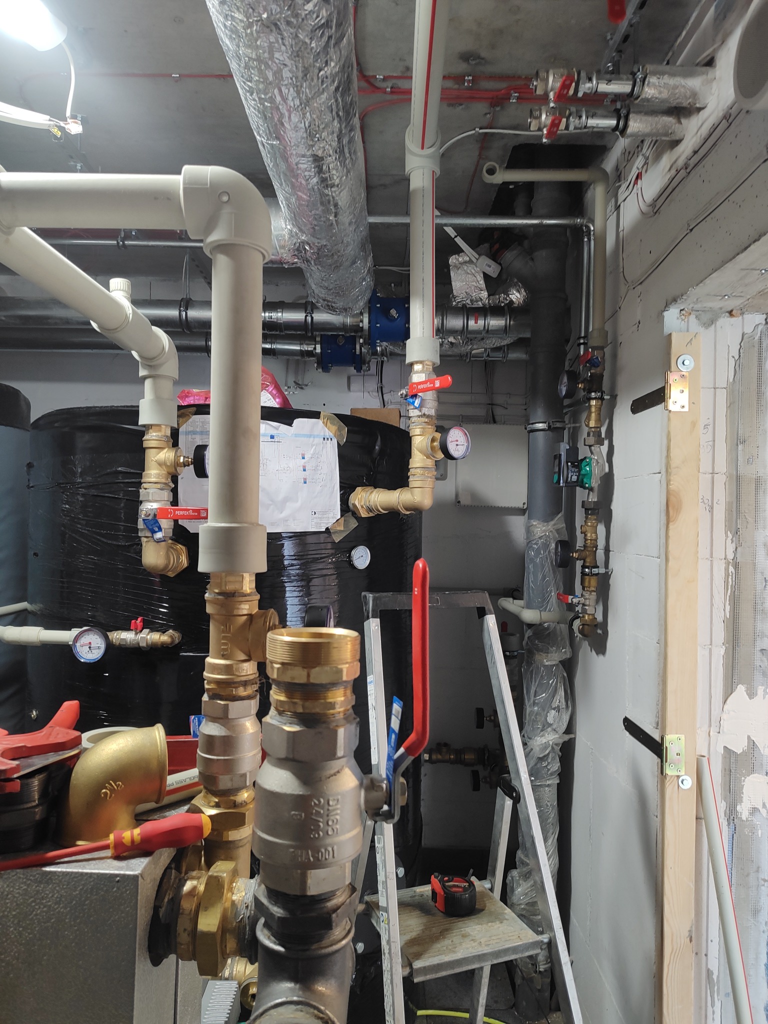 Złożona instalacja hydrauliczna z rurami, zaworami i manometrami w kotłowni. Widoczne elementy instalacji grzewczej i wodnej, drabina i narzędzia.