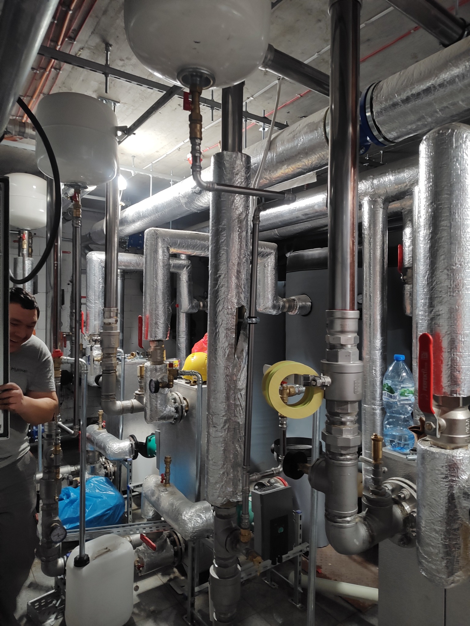 Złożona instalacja grzewcza z rurami w izolacji, zbiornikami wyrównawczymi i zaworami. Widoczny fragment osoby. Elementy instalacji podwieszone pod sufitem.