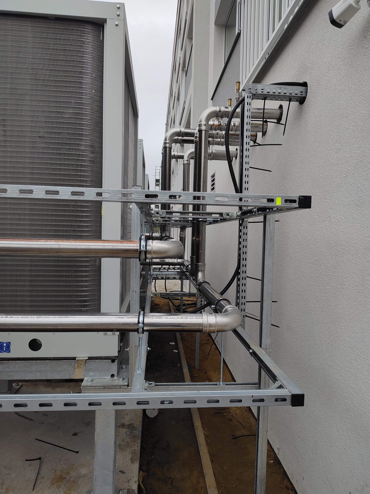 Zewnętrzna instalacja chłodnicza z rurami ze stali nierdzewnej, zamocowana do ściany budynku na metalowej konstrukcji wsporczej. Widoczne elementy wentylacyjne i zabezpieczenia.