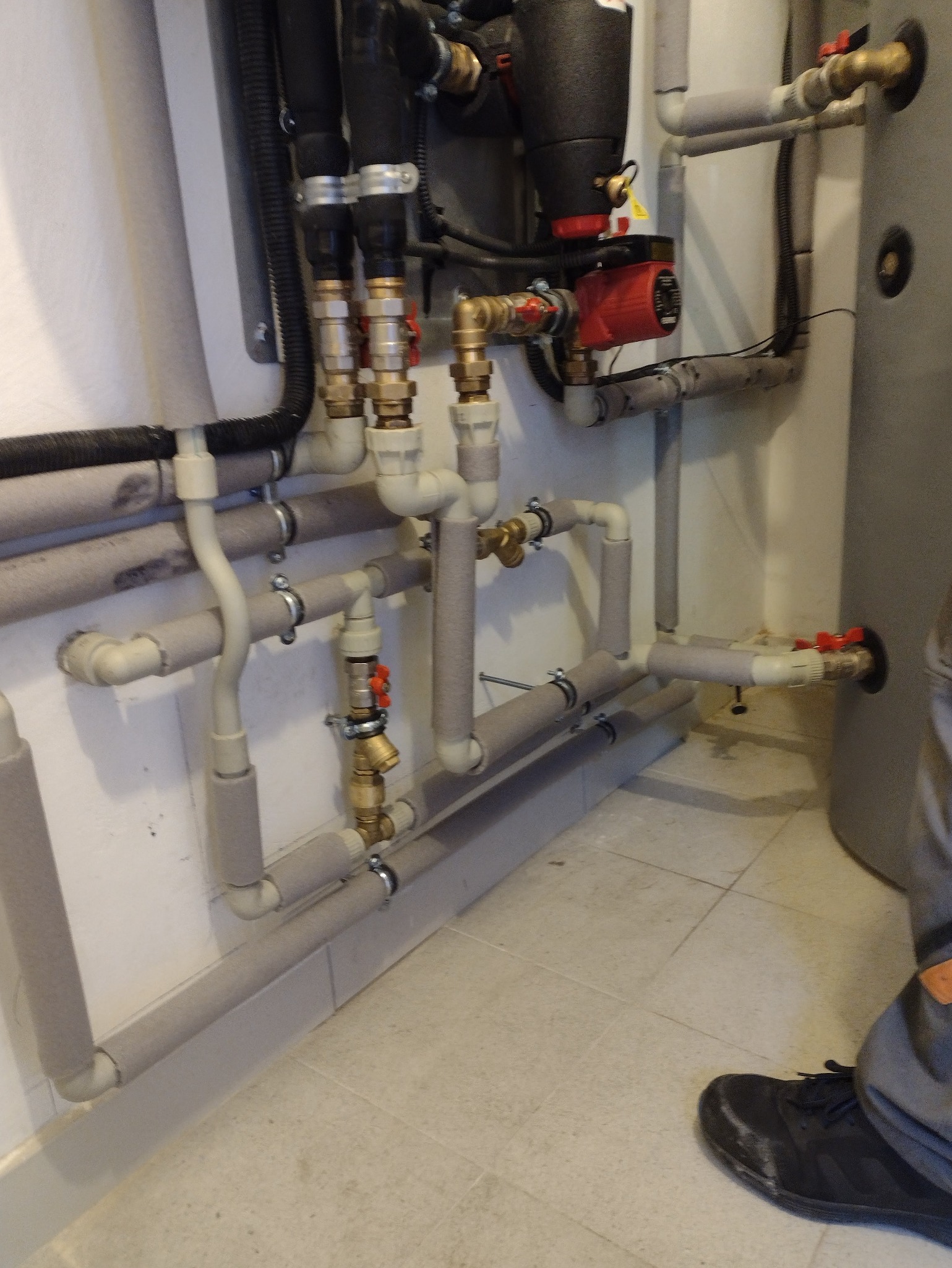 Złożona instalacja grzewcza z rurami w izolacji, zaworami i pompą obiegową, zamontowana przy ścianie i zbiorniku. Widoczny fragment osoby w dolnym rogu.