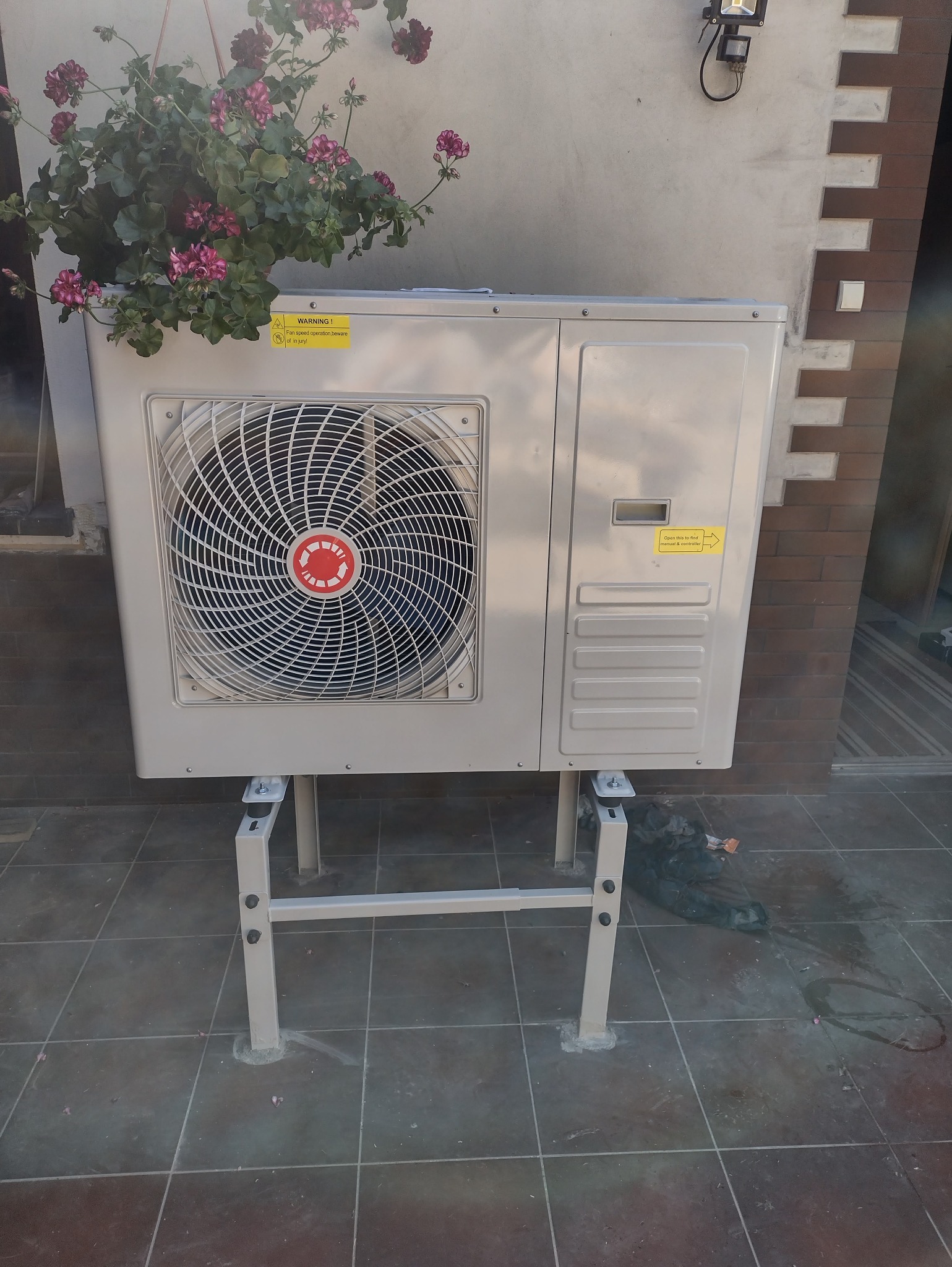 Pompa ciepła powietrze-woda na zewnątrz budynku, zamontowana na metalowym stelażu, z wiszącą doniczką z kwiatami nad nią. Widoczne płytki na podłożu.