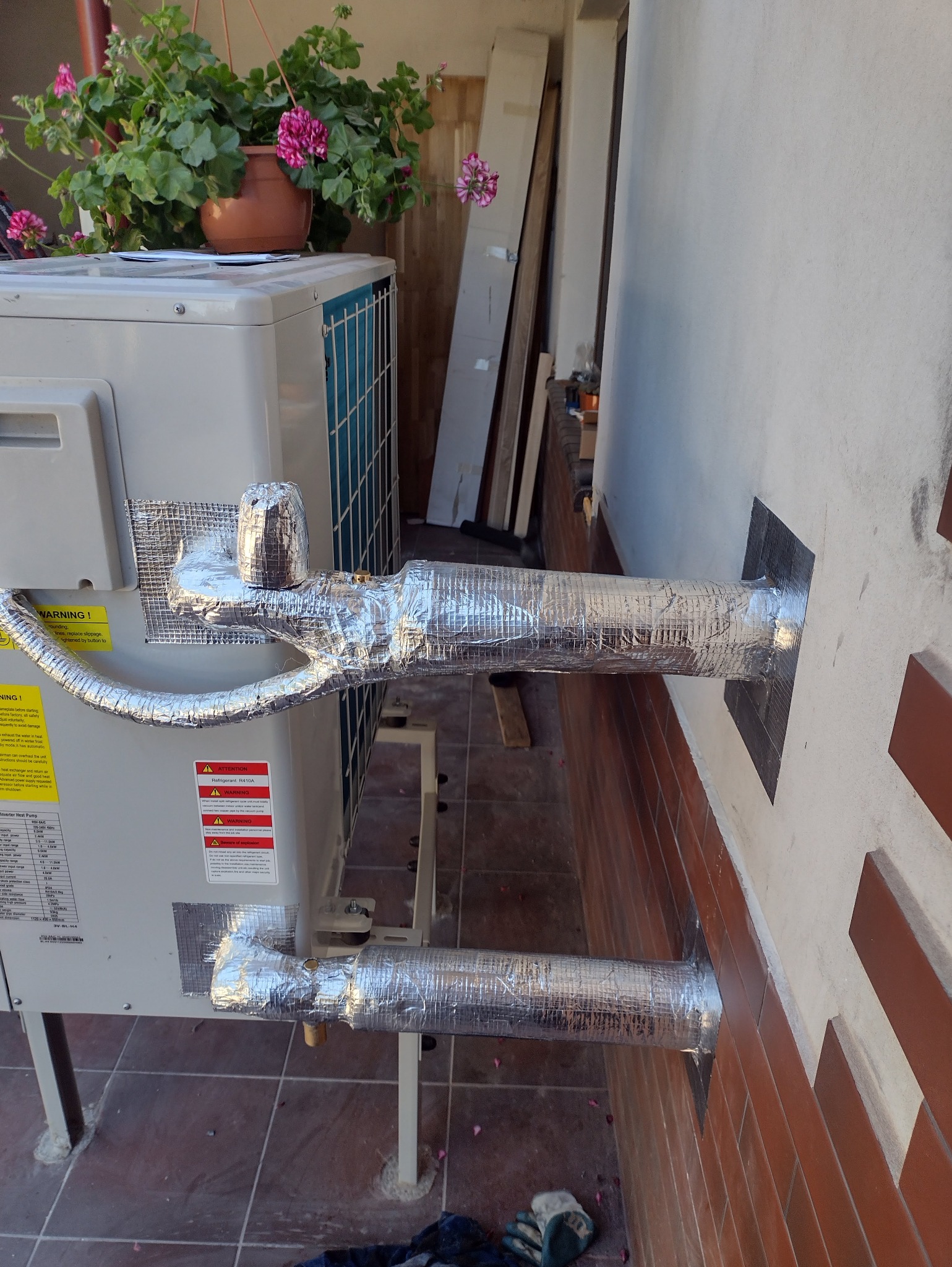 Zewnętrzna jednostka klimatyzacji z izolowanymi rurami, przymocowanymi do ściany budynku. Na górze jednostki doniczka z kwitnącymi kwiatami.