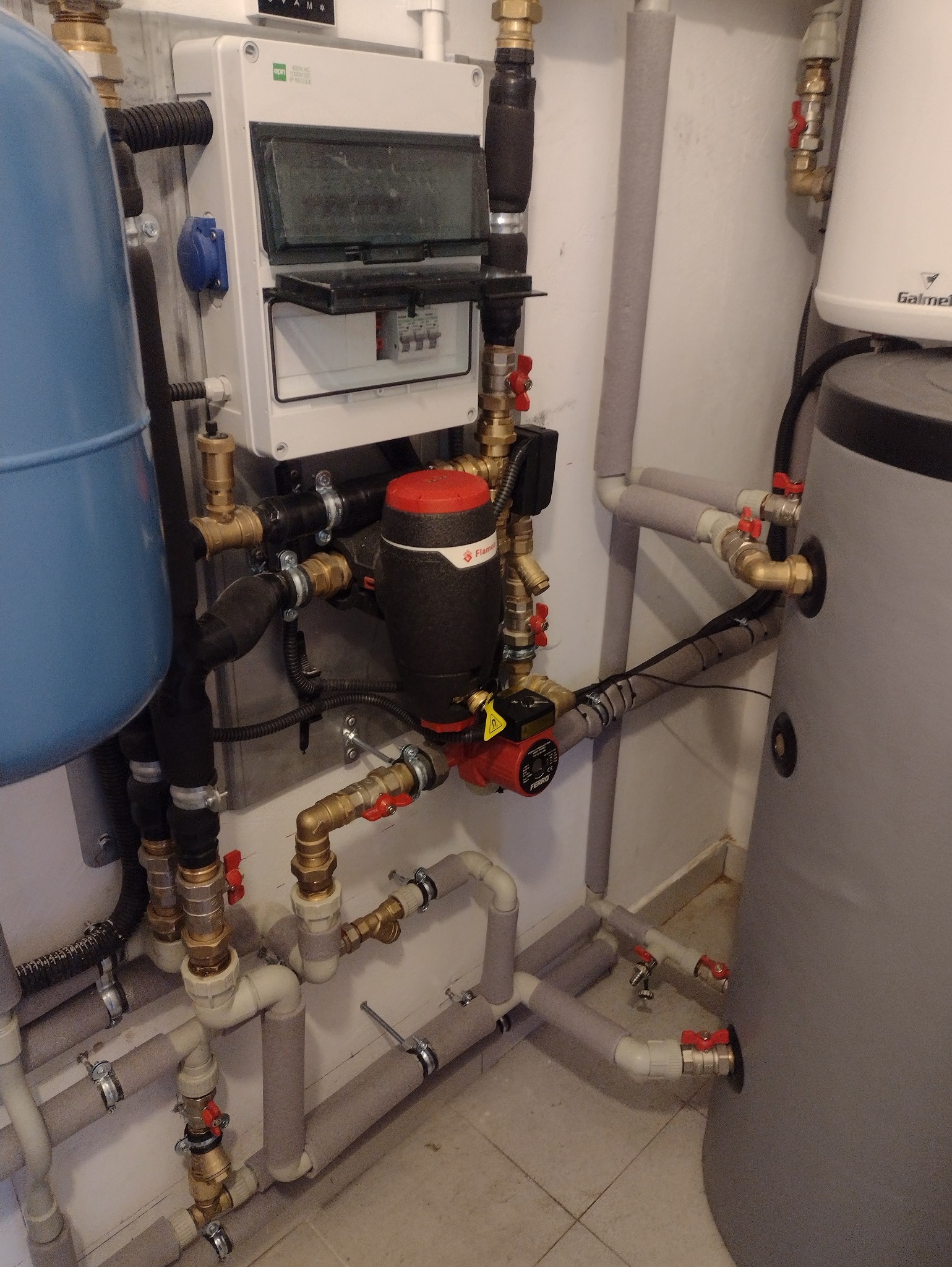 Węzeł cieplny z pompą obiegową, zaworami i zbiornikiem wyrównawczym. Widoczna instalacja z rur w izolacji i skrzynka elektryczna.