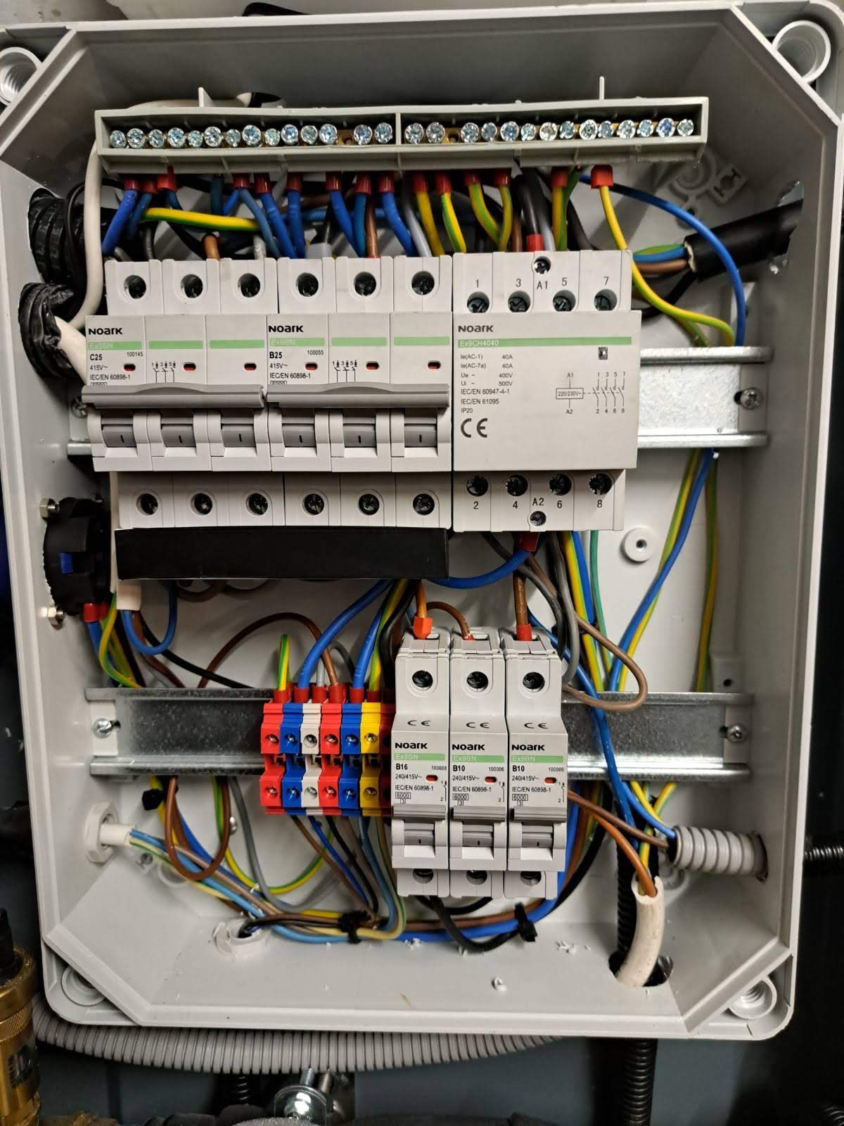 Rozdzielnica elektryczna w obudowie, widoczne bezpieczniki, przewody w różnych kolorach (niebieski, żółto-zielony, brązowy), listwy zaciskowe i szyny montażowe.