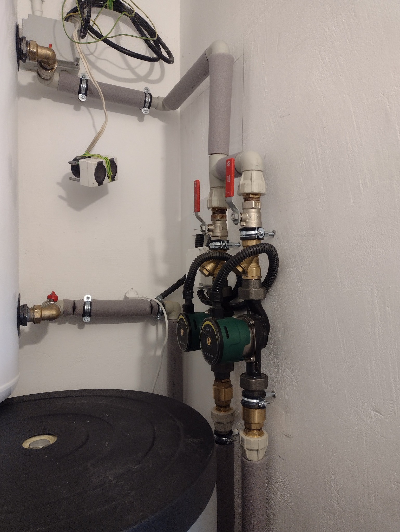 Szczegółowe ujęcie instalacji grzewczej z dwoma pompami obiegowymi, zaworami z czerwonymi uchwytami i izolowanymi rurami na tle białej ściany.