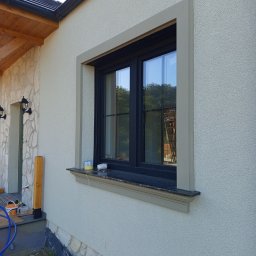 Decor-Jacks - Jasna elewacja domu z oknem w czarnej ramie, ozdobiona szarą sztukaterią. Widoczne narzędzia i materiały budowlane sugerują prace wykończeniowe.