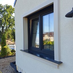 Decor-Jacks - Okna z czarną ramą i beżową listwą na elewacji budynku z jasnym tynkiem strukturalnym. W oknie odbija się budynek. Widoczny fragment ogrodu i lampa.