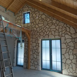 Decor-Jacks - Wnętrze z wykończoną ścianą z kamienia dekoracyjnego, drewnianym stropem i oknami zabezpieczonymi folią. Widoczna drabina i przygotowania do dalszych prac.