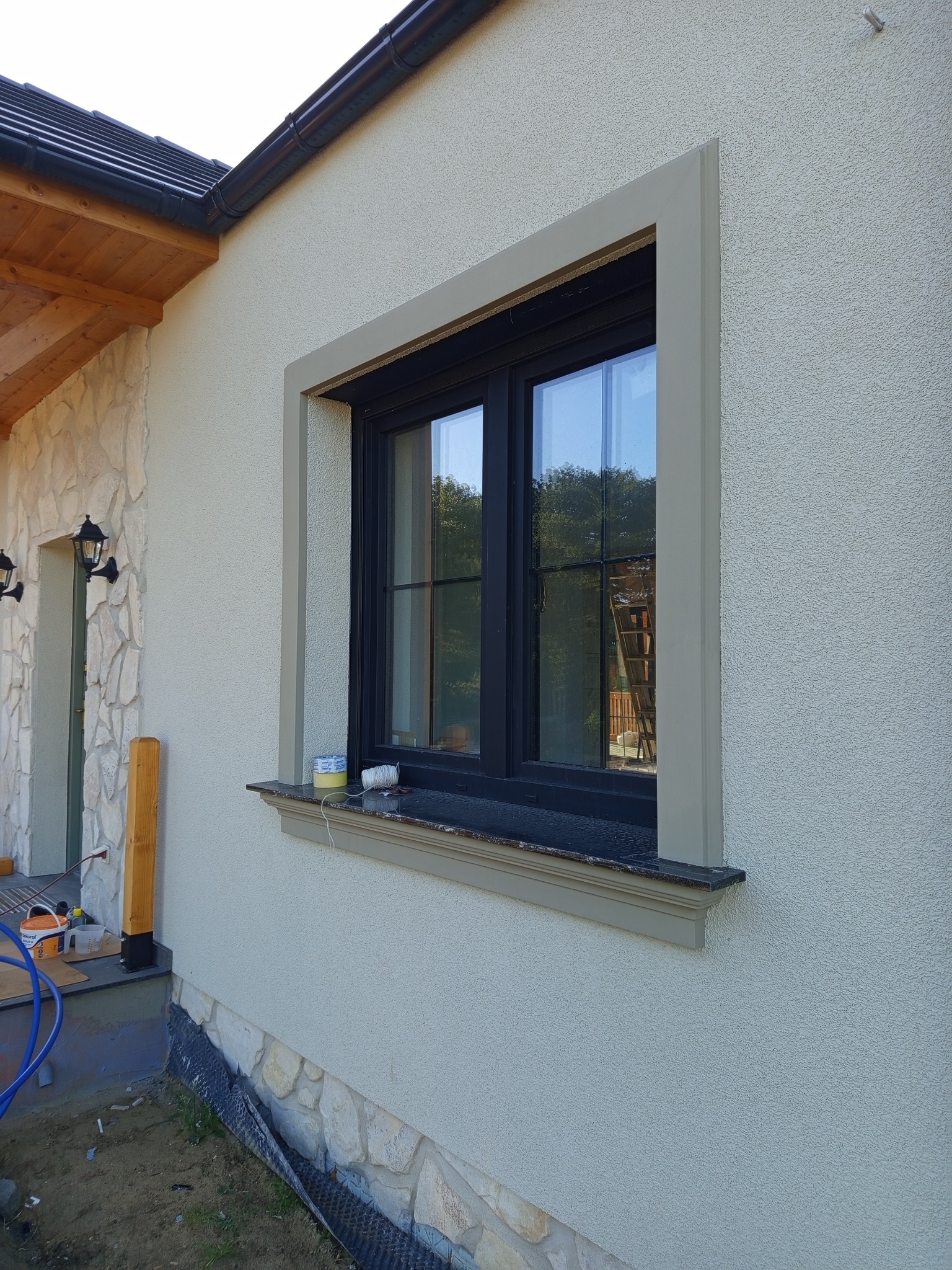 Jasna elewacja domu z oknem w czarnej ramie, ozdobiona szarą sztukaterią. Widoczne narzędzia i materiały budowlane sugerują prace wykończeniowe.
