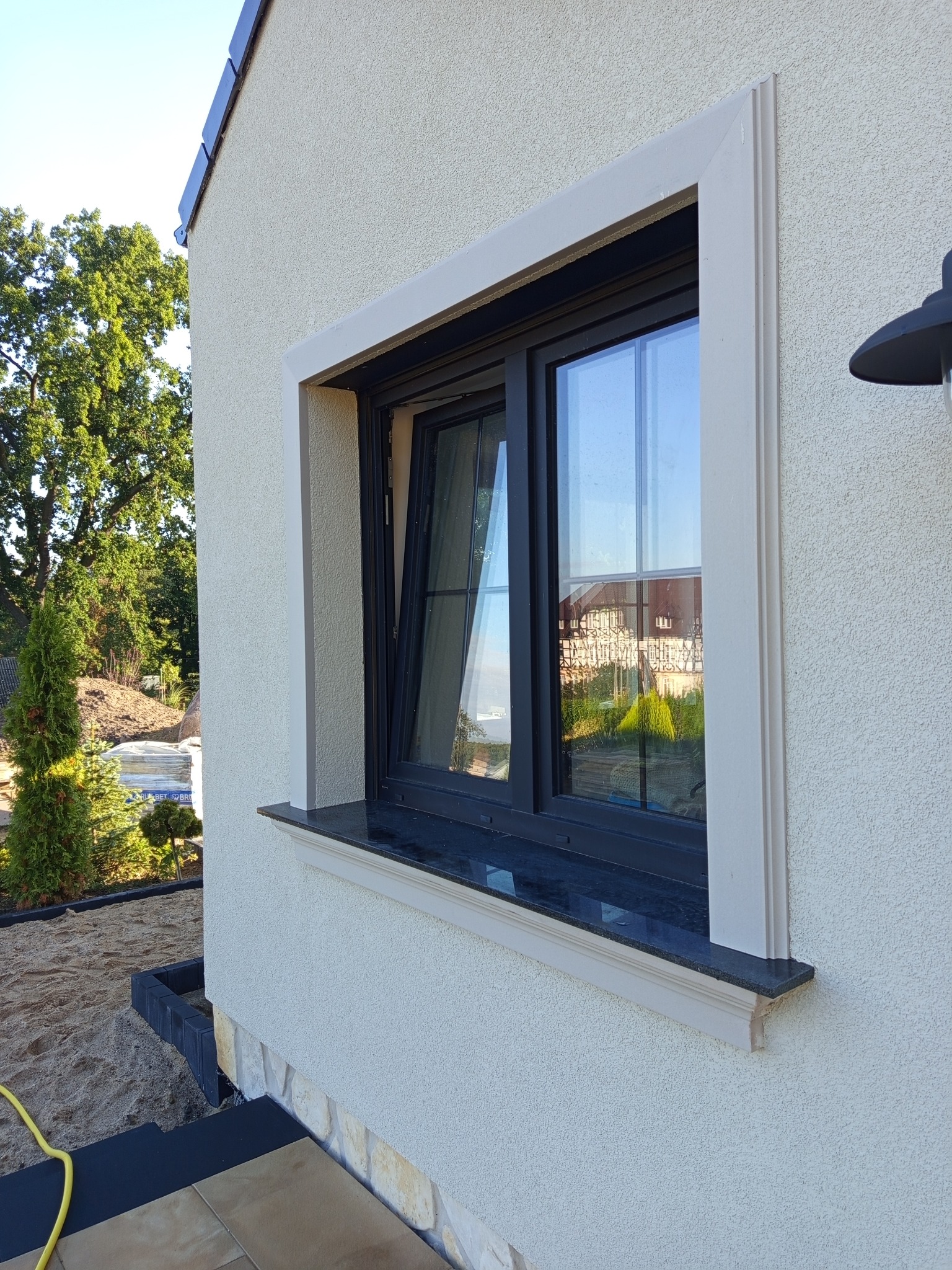 Okna z czarną ramą i beżową listwą na elewacji budynku z jasnym tynkiem strukturalnym. W oknie odbija się budynek. Widoczny fragment ogrodu i lampa.