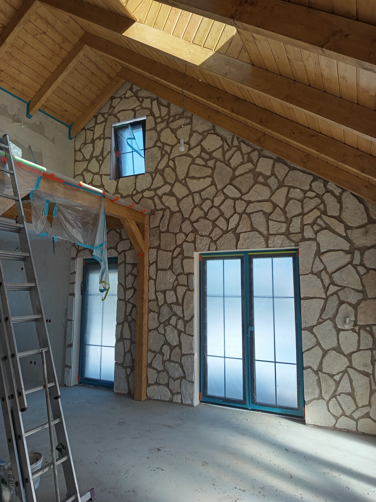 Wnętrze z wykończoną ścianą z kamienia dekoracyjnego, drewnianym stropem i oknami zabezpieczonymi folią. Widoczna drabina i przygotowania do dalszych prac.