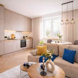 Studio wnętrz - Jasne, beżowe wnętrze mieszkania z aneksem kuchennym: zabudowa kuchenna, drewniany parkiet, sofa z poduszkami i okrągły stół. Nowoczesny design, ciepłe oświetlenie.