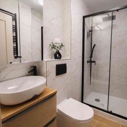 Studio wnętrz - Nowoczesna łazienka z kabiną prysznicową typu walk-in, umywalką nablatową na drewnianej szafce i marmurowymi płytkami ściennymi z delikatnym złotym żyłkowaniem.