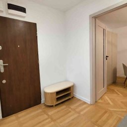 Studio wnętrz - Jasne wnętrze mieszkania po remoncie: drewniana podłoga w jodełkę, białe ściany, brązowe drzwi wejściowe i beżowa szafka na buty. Widok na jadalnię przez otwarte drzwi.