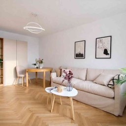 Studio wnętrz - Jasne wnętrze salonu po odświeżeniu: białe ściany, parkiet w jodełkę, beżowa sofa i minimalistyczne dekoracje. Widoczne obrazy na ścianie i nowoczesna lampa.