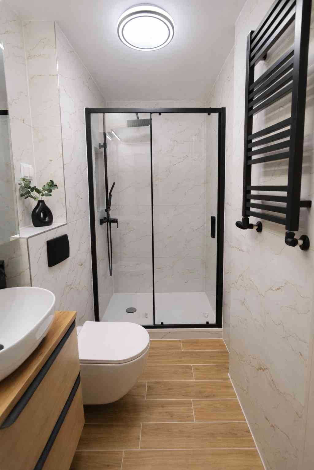 Nowoczesna łazienka z kabiną prysznicową w czarnej ramie, drewnopodobną podłogą i marmurowymi płytkami. Czarny grzejnik drabinkowy i umywalka na drewnianej szafce.