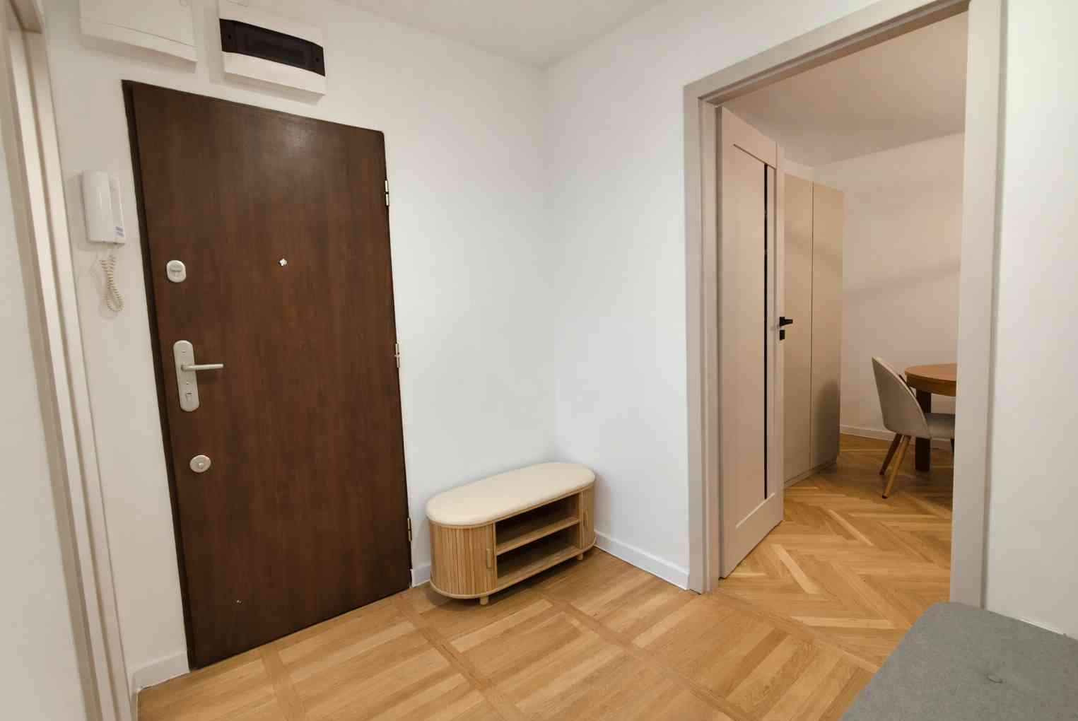 Jasne wnętrze mieszkania po remoncie: drewniana podłoga w jodełkę, białe ściany, brązowe drzwi wejściowe i beżowa szafka na buty. Widok na jadalnię przez otwarte drzwi.