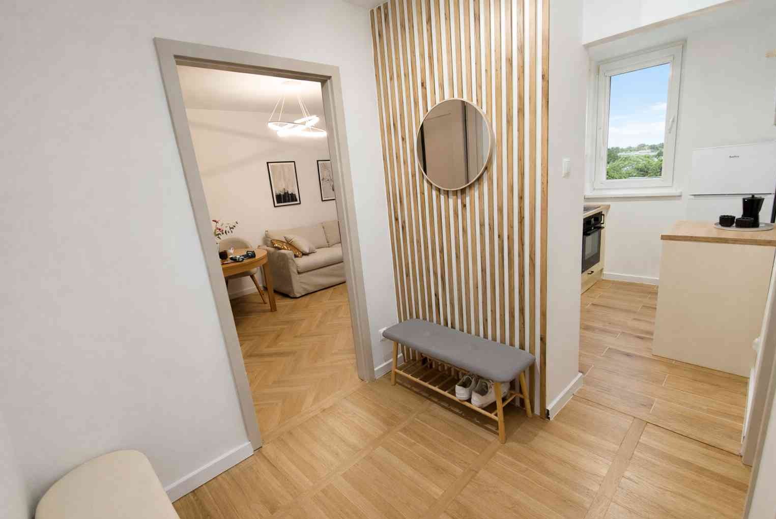 Nowoczesny przedpokój po remoncie: drewniane lamele z okrągłym lustrem, szary tapicerowany siedzisko, podłoga z płytek imitujących drewno, widok na salon i kuchnię.