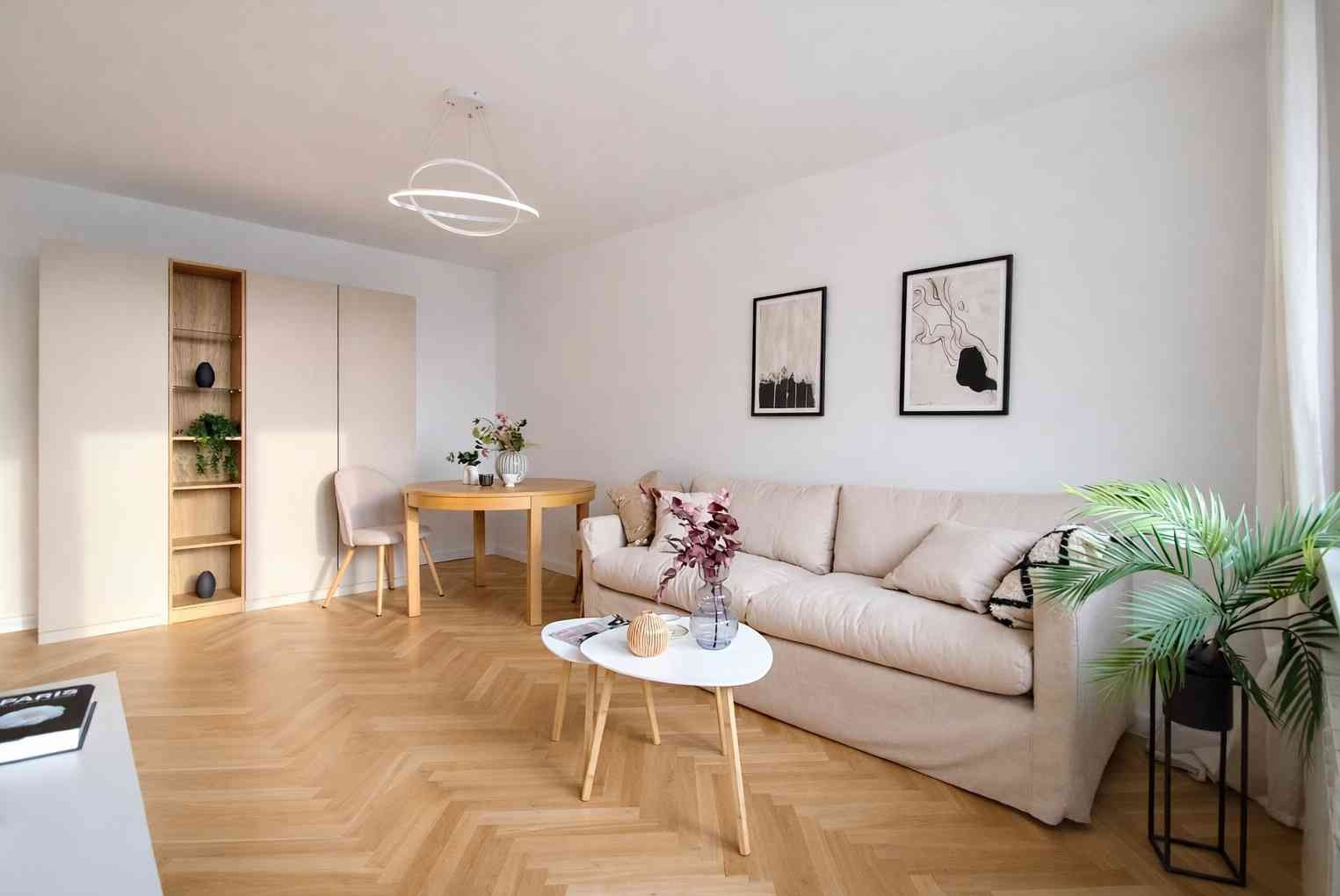 Jasne wnętrze salonu po odświeżeniu: białe ściany, parkiet w jodełkę, beżowa sofa i minimalistyczne dekoracje. Widoczne obrazy na ścianie i nowoczesna lampa.