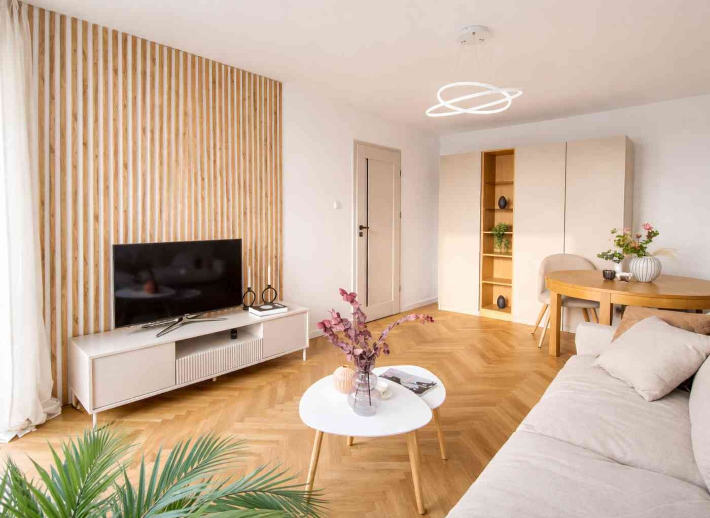 Jasne wnętrze salonu z drewnianą ścianą z listew, telewizorem na szafce RTV i parkietem w jodełkę. Minimalistyczny design z roślinami i dekoracjami.