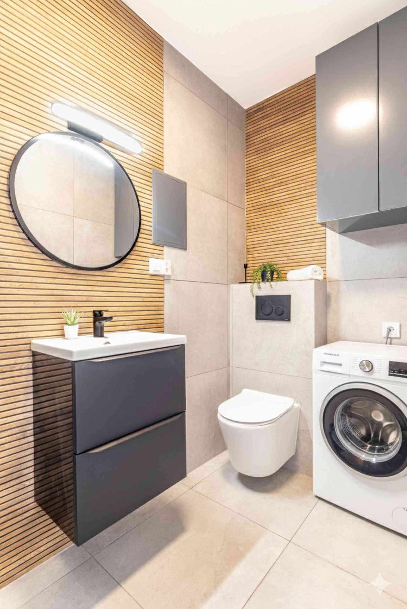 Nowoczesna łazienka z drewnianą boazerią, okrągłym lustrem z podświetleniem, szarą szafką pod umywalką i pralką. Minimalistyczny design, jasne płytki podłogowe.