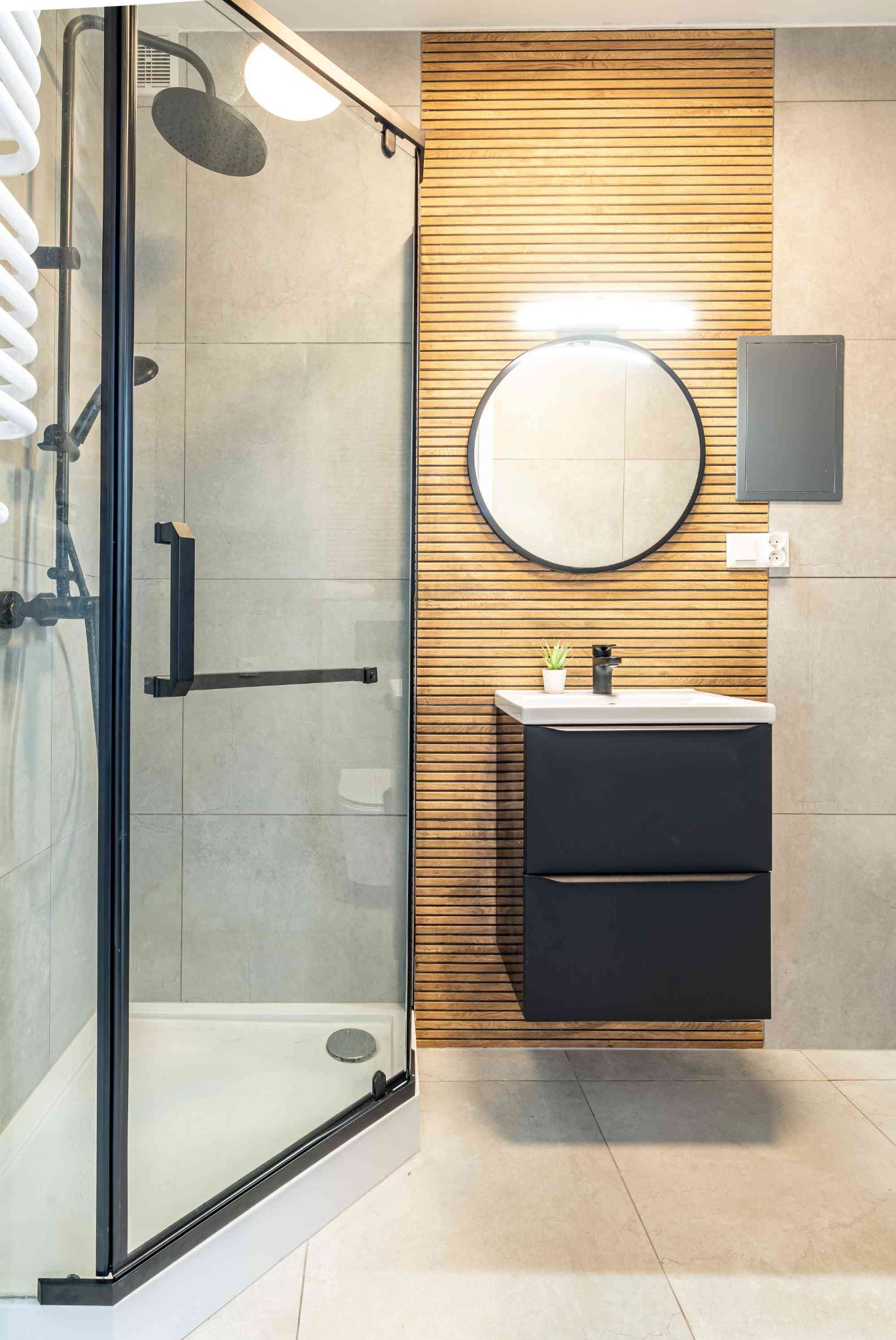 Nowoczesna łazienka z kabiną prysznicową typu walk-in, umywalką z szafką w kolorze grafitowym i drewnianą ścianą z lamelami. Minimalistyczny design, jasne płytki.