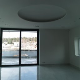 Wojtholm Bygg - Jasne wnętrze salonu z dużym oknem balkonowym i widokiem na jezioro. Na suficie okrągła dekoracja. Podłoga wyłożona błyszczącymi, jasnymi płytkami.