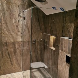 Wojtholm Bygg - Nowoczesna łazienka z kabiną prysznicową typu walk-in, wykończona płytkami imitującymi marmur w odcieniach brązu. Widoczne detale: półka w ścianie, bateria prysznicowa.