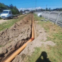 Cyngielkop usługi minikoparka - Pomarańczowe rury drenażowe ułożone w wykopie, widok z góry. W tle biały samochód dostawczy i dom. Słoneczny dzień, niebieskie niebo.