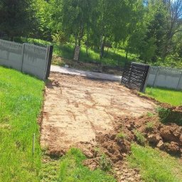 Cyngielkop usługi minikoparka - Przygotowany teren pod budowę, wyrównana ziemia, widoczne ślady opon, ogrodzenie betonowe i brama w tle, zieleń. Teren przygotowany pod inwestycję.