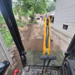 Cyngielkop usługi minikoparka - Widok z kabiny koparki na wykop wzdłuż budynku. Żółta maszyna w trakcie pracy, ziemia świeżo naruszona, widoczne elementy sterowania koparki.