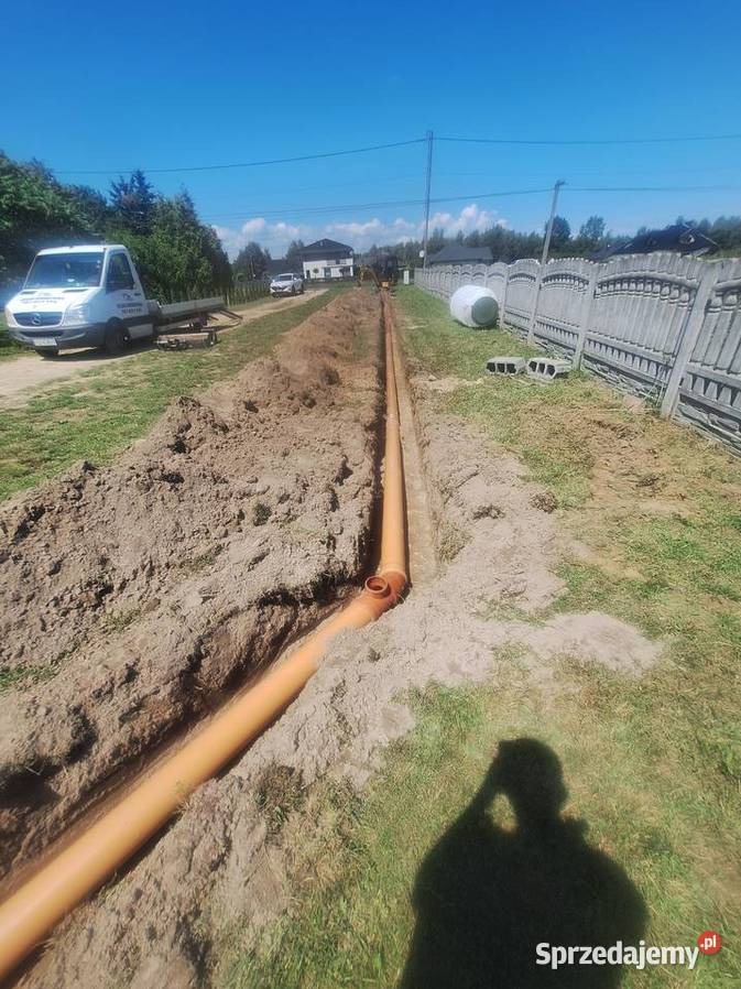 Pomarańczowe rury drenażowe ułożone w wykopie, widok z góry. W tle biały samochód dostawczy i dom. Słoneczny dzień, niebieskie niebo.