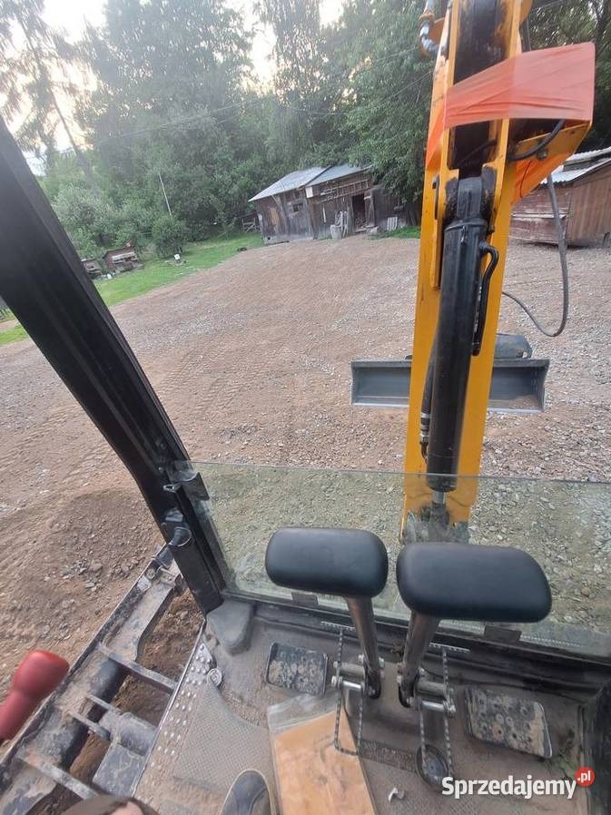 Widok z kabiny żółtej mini koparki na teren z budynkiem gospodarczym w tle. Dźwignie sterujące i szyba kabiny w pierwszym planie. Lekkie zabrudzenia na podłodze.