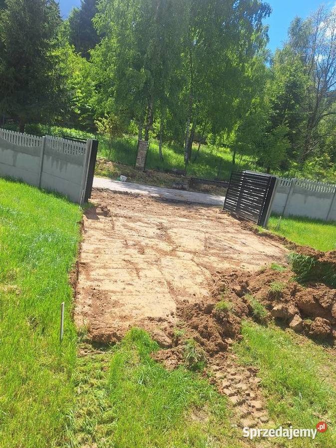 Przygotowany teren pod budowę, wyrównana ziemia, widoczne ślady opon, ogrodzenie betonowe i brama w tle, zieleń. Teren przygotowany pod inwestycję.