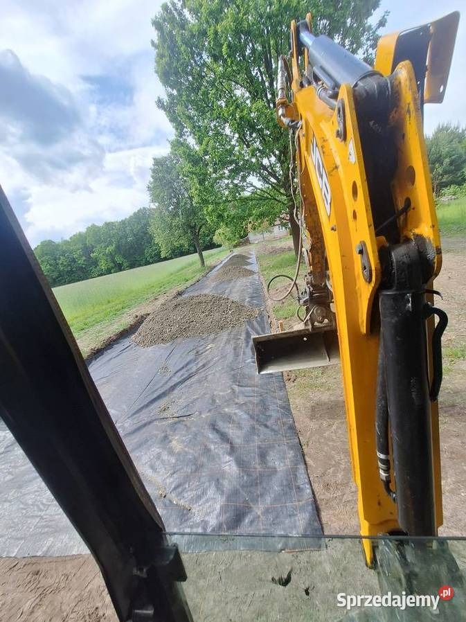 Żółta koparka na czarnej geowłókninie z nasypanym żwirem, widok z kabiny operatora, w tle zieleń drzew i łąka. Prace przygotowawcze pod budowę.
