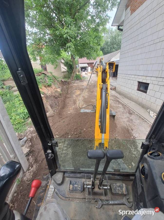 Widok z kabiny koparki na wykop wzdłuż budynku. Żółta maszyna w trakcie pracy, ziemia świeżo naruszona, widoczne elementy sterowania koparki.