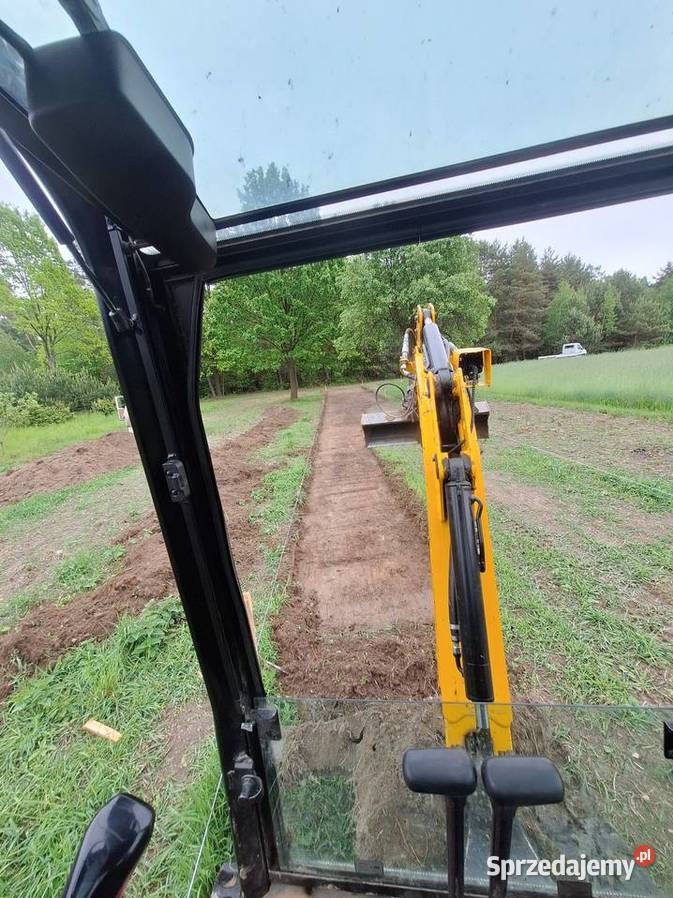 Widok z kabiny żółtej koparki podczas wykonywania wykopu w ziemi na zewnątrz, w tle zieleń i drzewa. Praca w toku, wąski wykop.