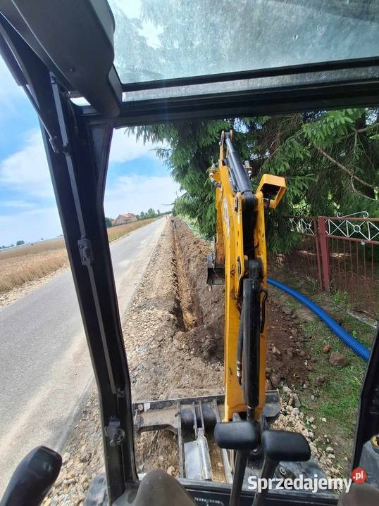 Żółta koparka z kabiny operatora, wykonująca wykop wzdłuż drogi. Widoczny rów, niebieski wąż i fragment krajobrazu. Ujęcie z perspektywy pierwszej osoby.